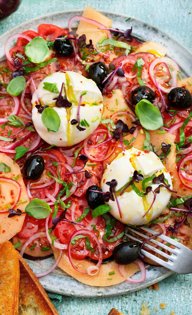 Eine Kugel Burrata auf einem Bett aus Melonen- und Tomatenscheiben getoppt von Zwiebeln und Oliven