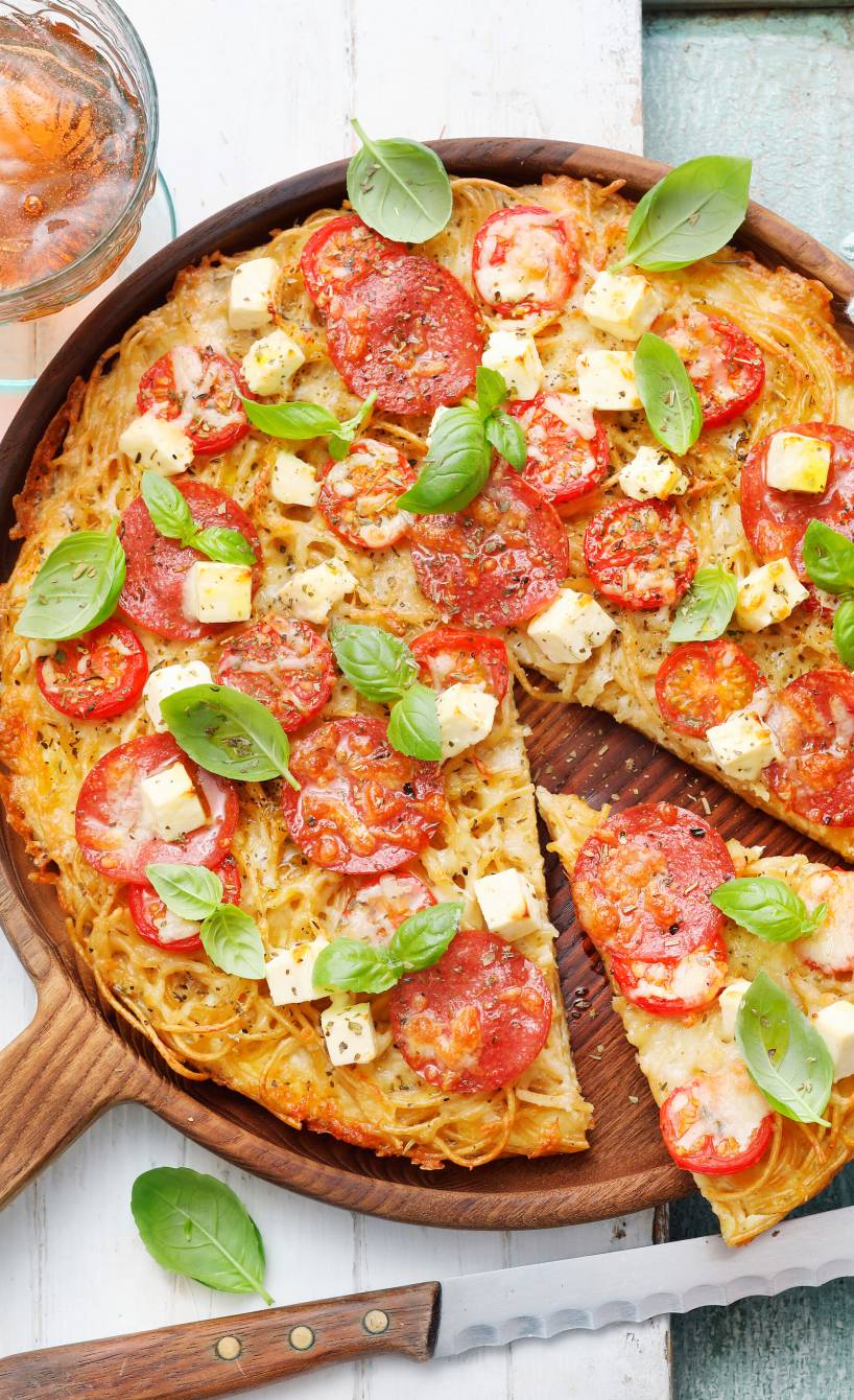 Runde Spaghettipizza mit Tomaten, Mozzarella und Feta angeschnitten auf einem Holzteller