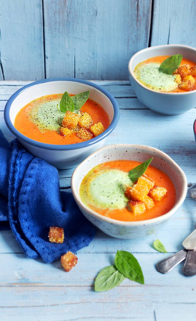 Tomatensuppe in mehreren Suppenschalen getoppt mit mit Basilikumschaum und Parmesan-Polenta-Croûtons auf einem Holztisch