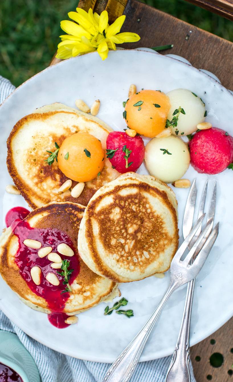 Pancakes auf einem Teller getoppt von Melonenkugeln und Himbeersauce auf einem Gartenstuhl