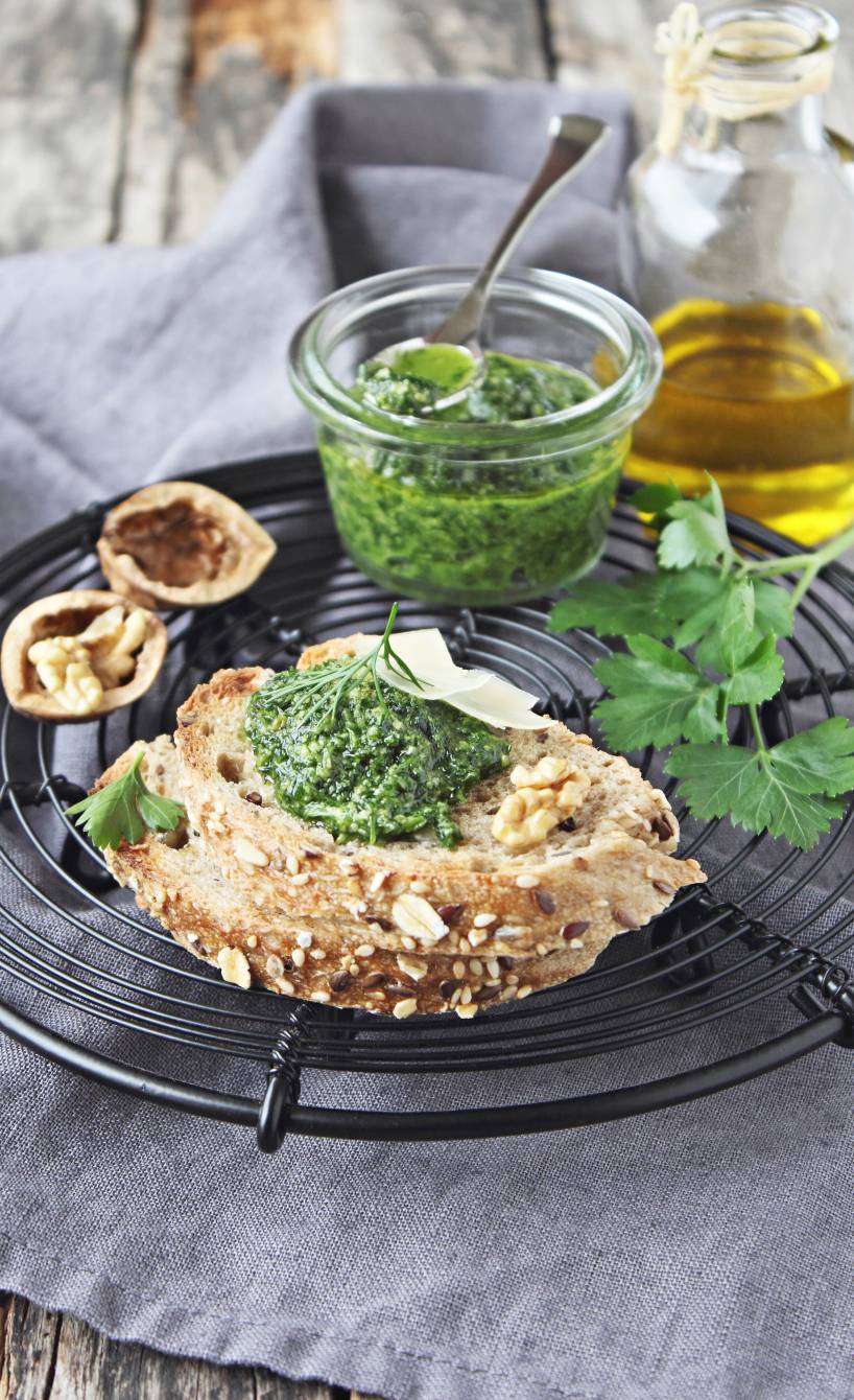 Grünes Pesto auf einer Scheibe Baguette, daneben Walnüsse und ein Glas mit Pesto auf einem Tisch