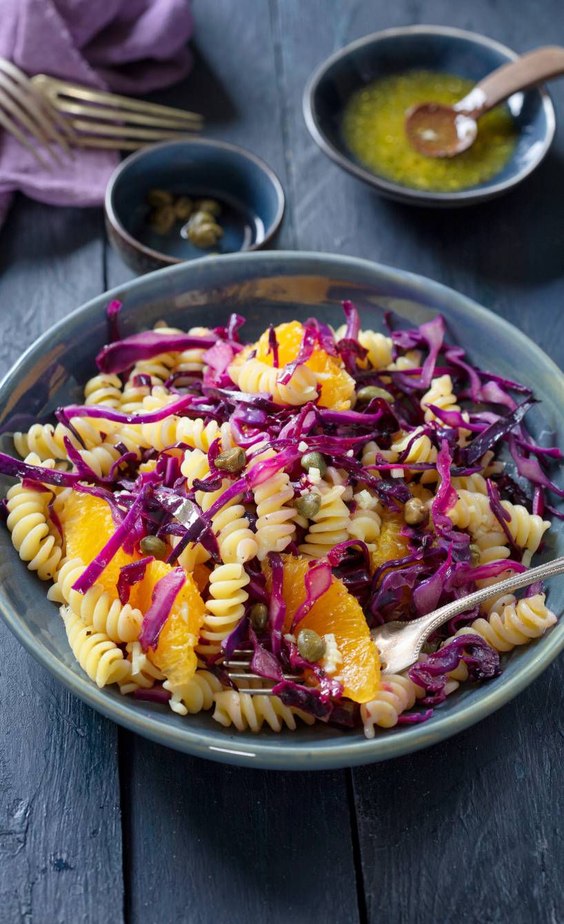 Nudelsalat mit Rotkohl, Kapern und Orangen angerichtet auf einem tiefen Teller, daneben Dressing in einer Schale