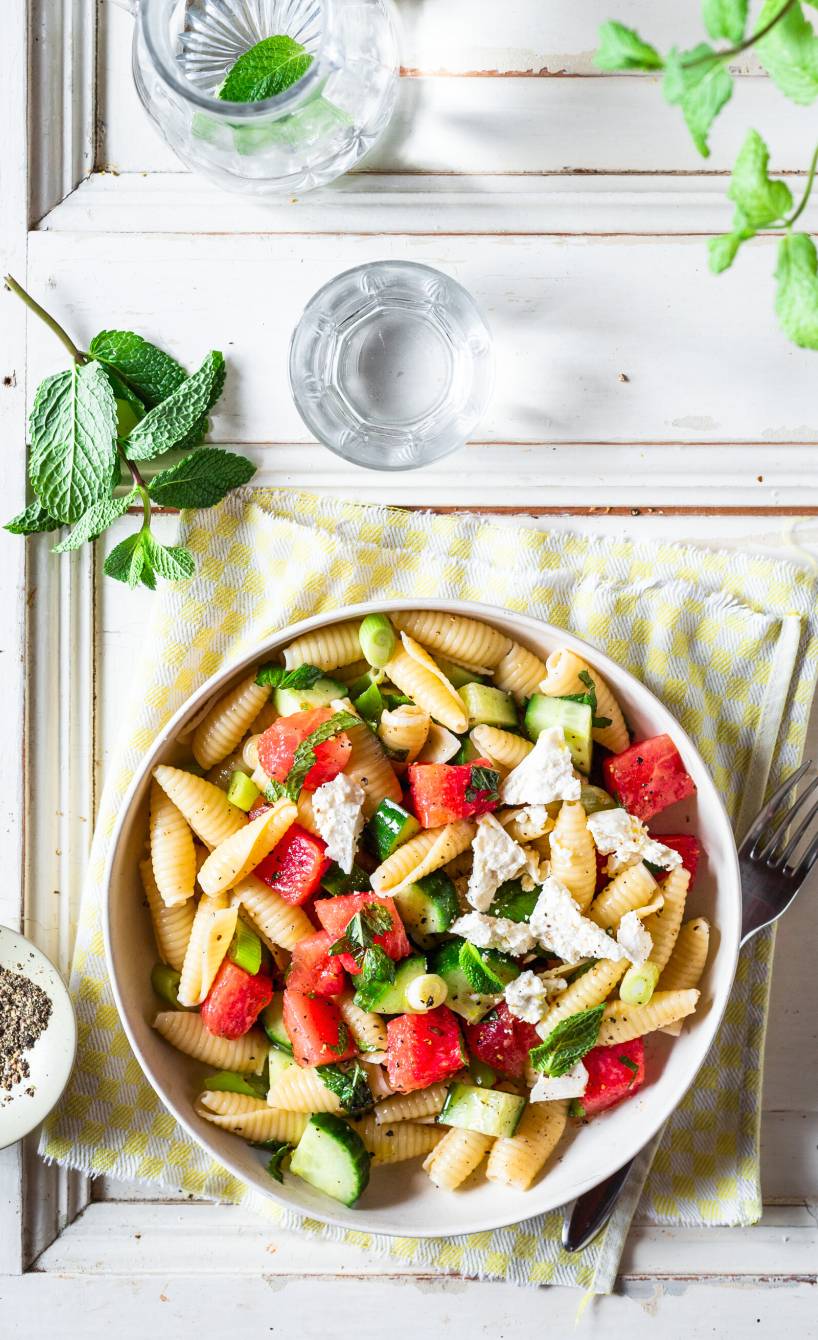 Pastasalat in einer Schüssel mit Wassermelonenstücken, Gurke, Fetawürfeln und Minzblättern auf einem Tisch