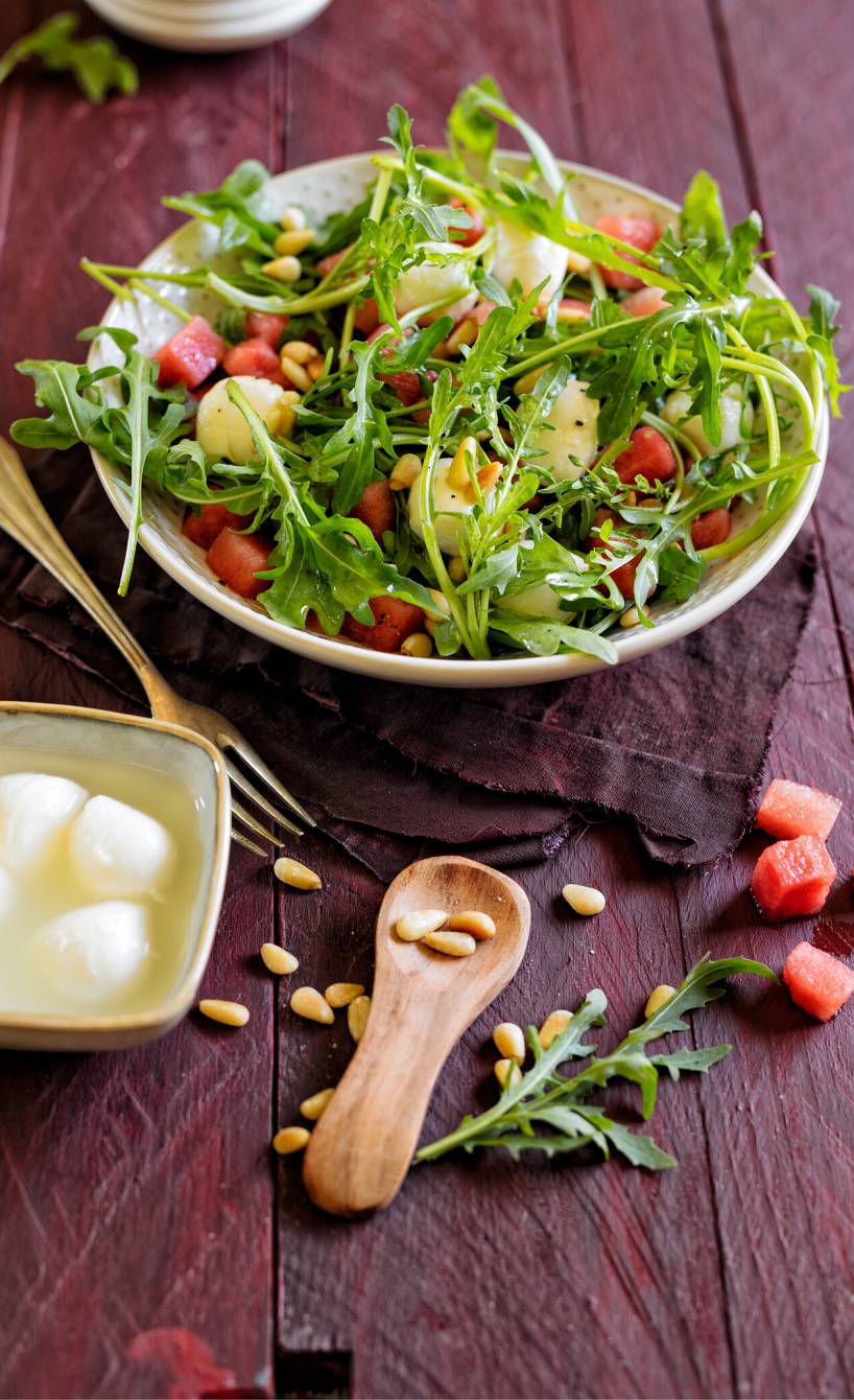 Wassermelonensalat mit Mini-Mozzarellakugeln und Rucola auf einem Teller, daneben Pinienkerne