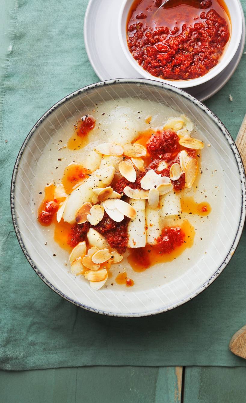 Weiße Spargelsuppe in einer Schale getoppt mit Tomatenöl und Mandelblättchen, daneben einige Scheiben Ciabatta