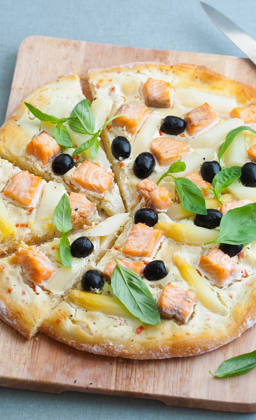 Runde Spargelpizza mit Lachs auf einem Holzbrett in Pizzastücke geschnitten