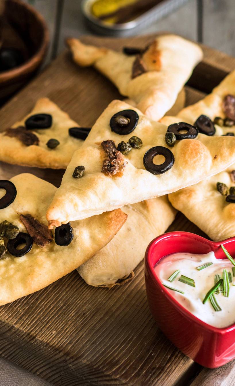 Pizzaecken mit Sardellen und Oliven auf einem Holzbrett, daneben Rosmarindip