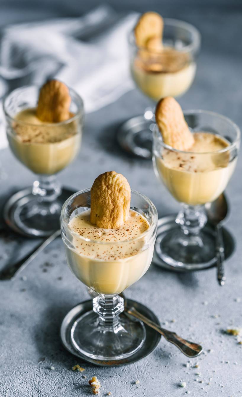 Zabaione in vier Gläsern getoppt von jeweils einem Löffelbiskuit und Marsala auf einem Tisch