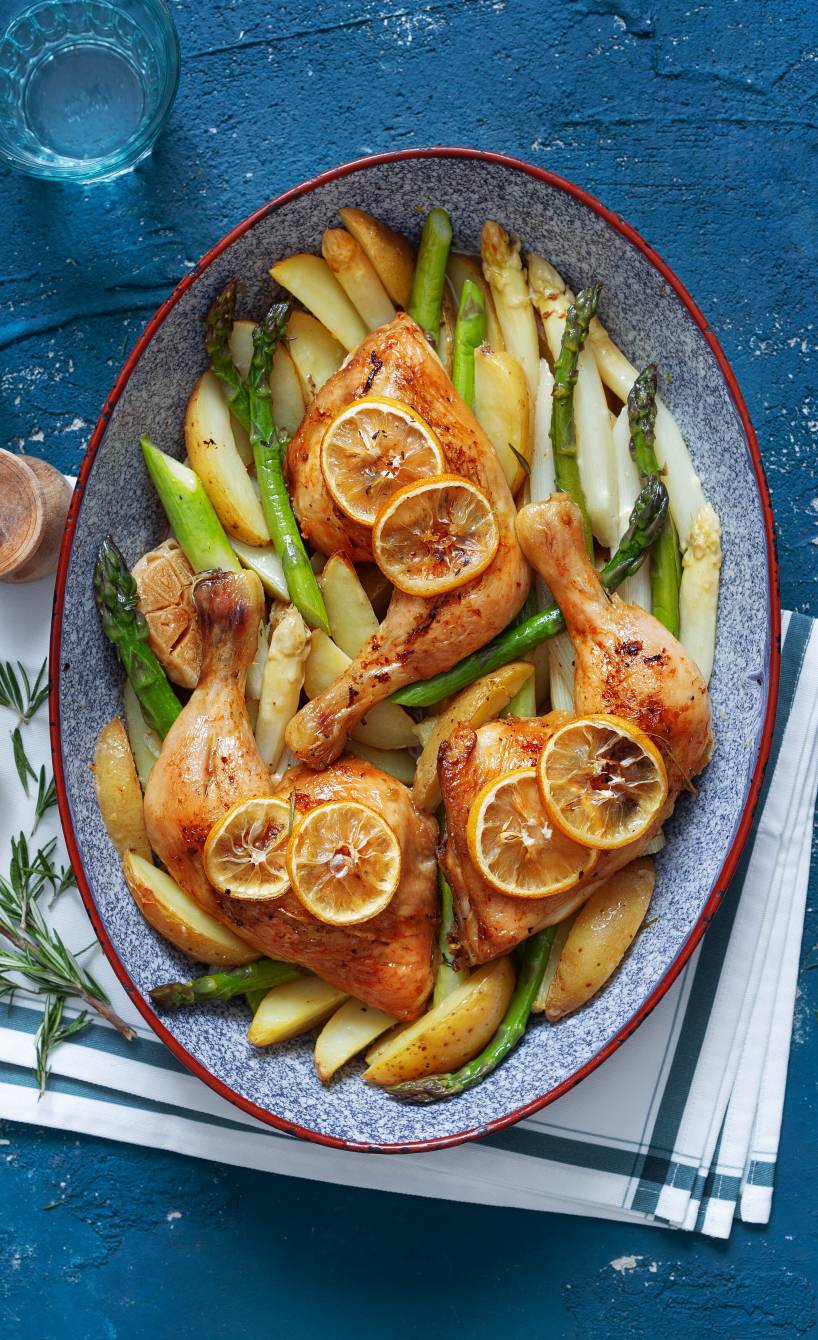 Gebackene Hähnchenkeulen mit Kartoffelspalten, grünem und weißem Spargel angerichtet auf einer Servierplatte