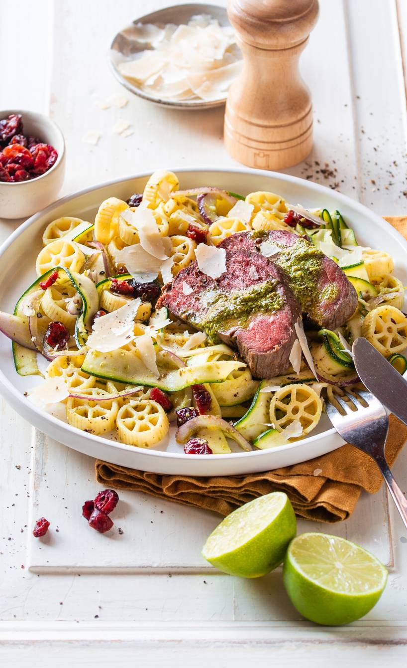 Italienischer Nudelsalat mit Zucchini angerichtet mit gebackenen Rinderfiletscheiben und grünem Pesto-Dressing auf einem Teller aus dem Backofen
