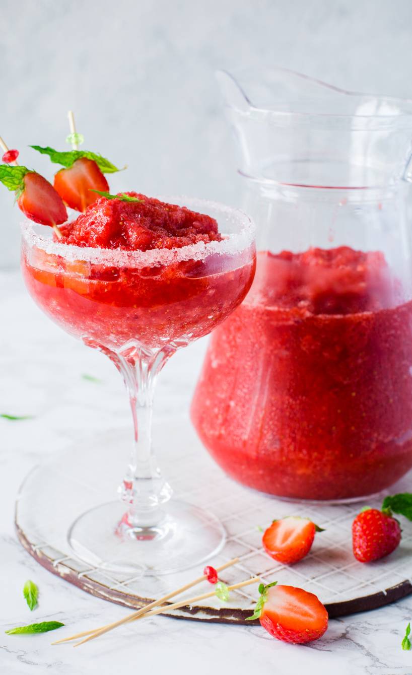 Selbst gemachter, dekorierter Strawberry Margarita in einem Coktailglas, dahinter eine Karaffe mit mehr