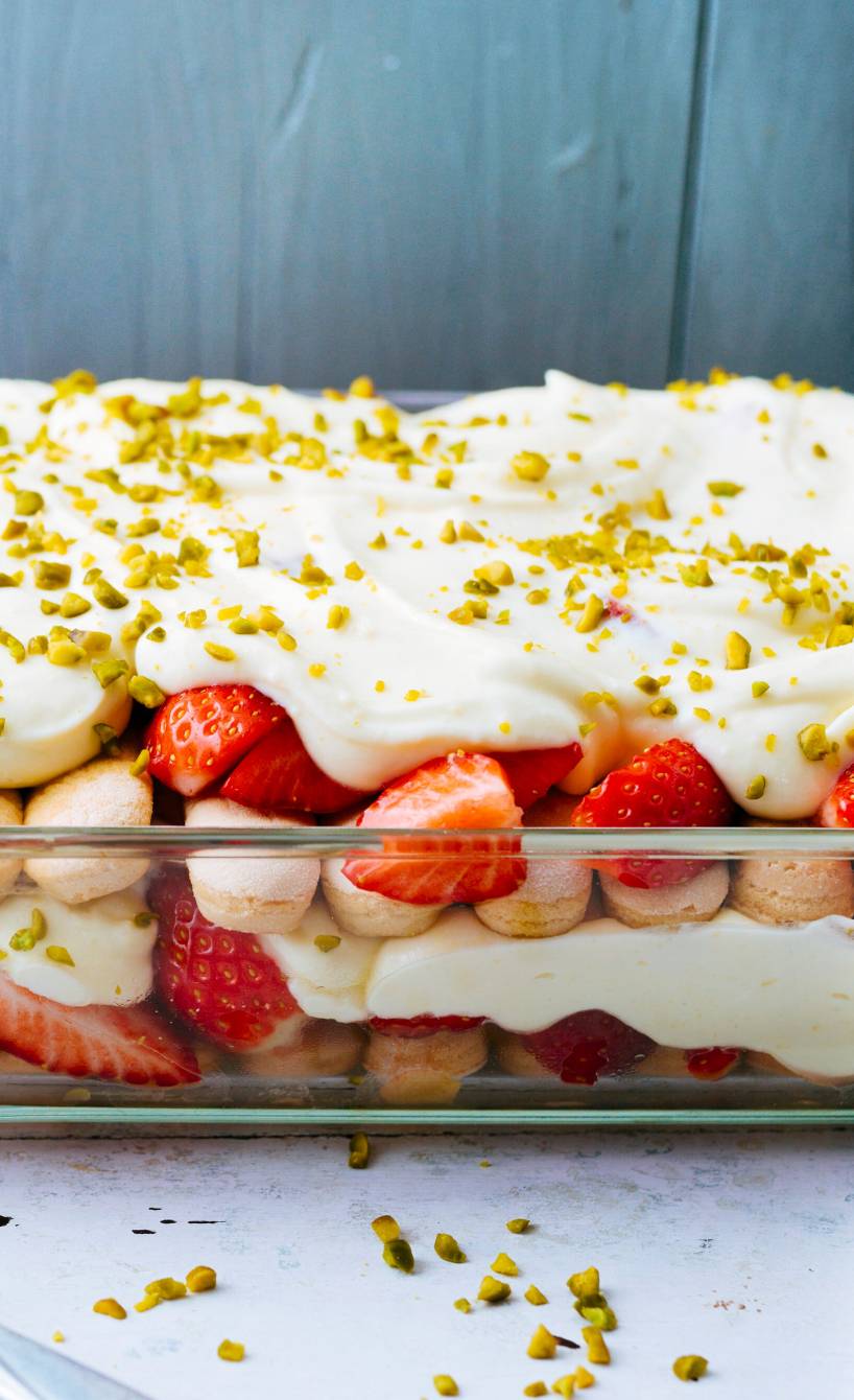 Erdbeertiramisu mit Quark und Joghurt in einer gläsernen Auflaufform bestreut mit Pistazien, daneben frische Erdbeeren