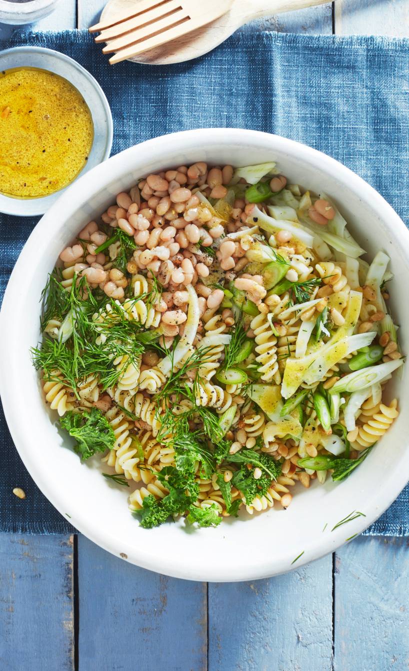Veganer italienischer Nudelsalat mit Fenchel, Grünkohl und Bohnen in einer Salatschüssel, daneben Pinienkerne und Dressing