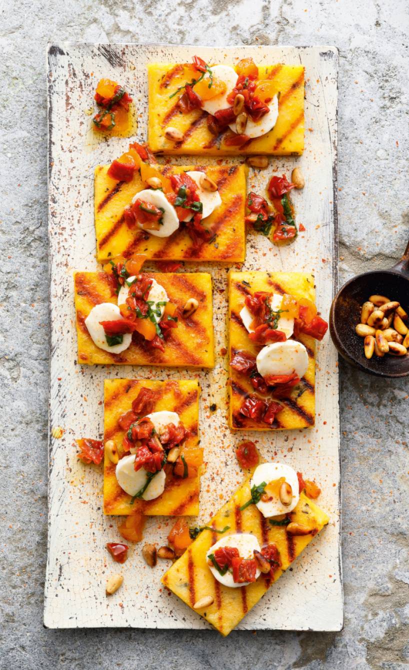 Polentaschnitten in Rechtecke geschnitten mit Grillstreifen getoppt von Tomaten, Mozzarella und Pinienkernen auf einem Teller