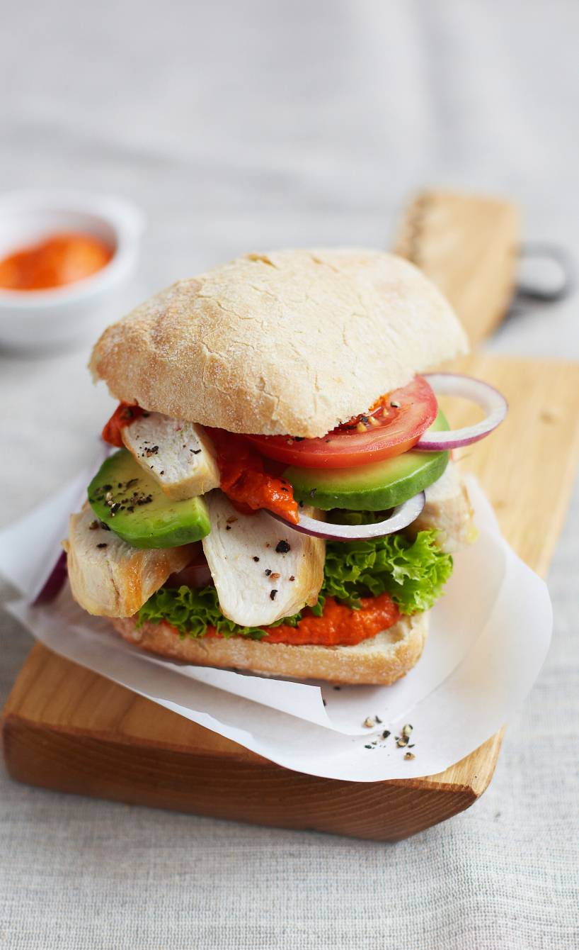 Ein Ciabatta-Burger belegt mit Hähnchenbrust, Avocado und pikanter Ajvar-Creme angerichtet auf einem Holzbrett