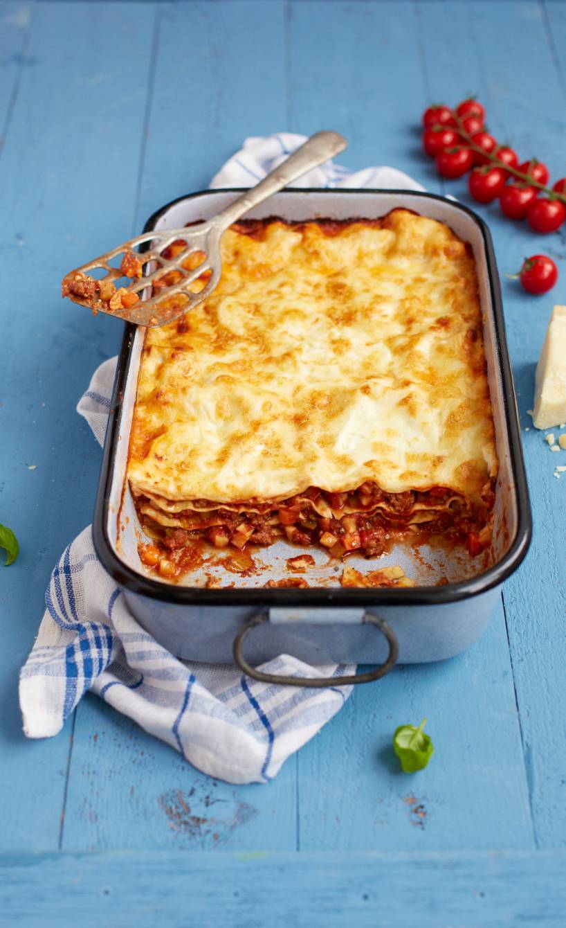 Lasagne mit Bolognese-Schichten in einer Auflaufform angeschnitten auf einem Tisch, daneben Parmesan und Tomaten