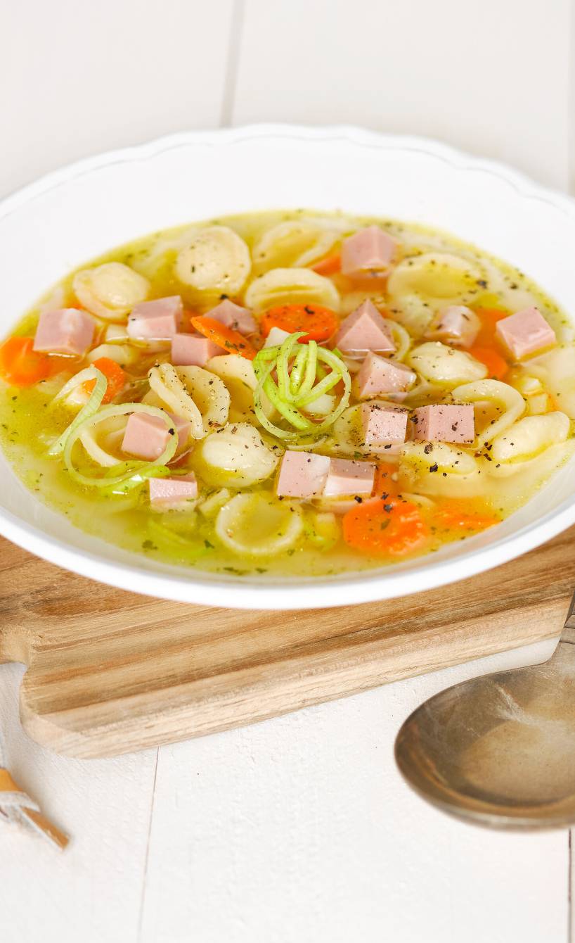 Nudelsuppe in einem Teller auf einem Holzbrett, daneben Mortadellastücke