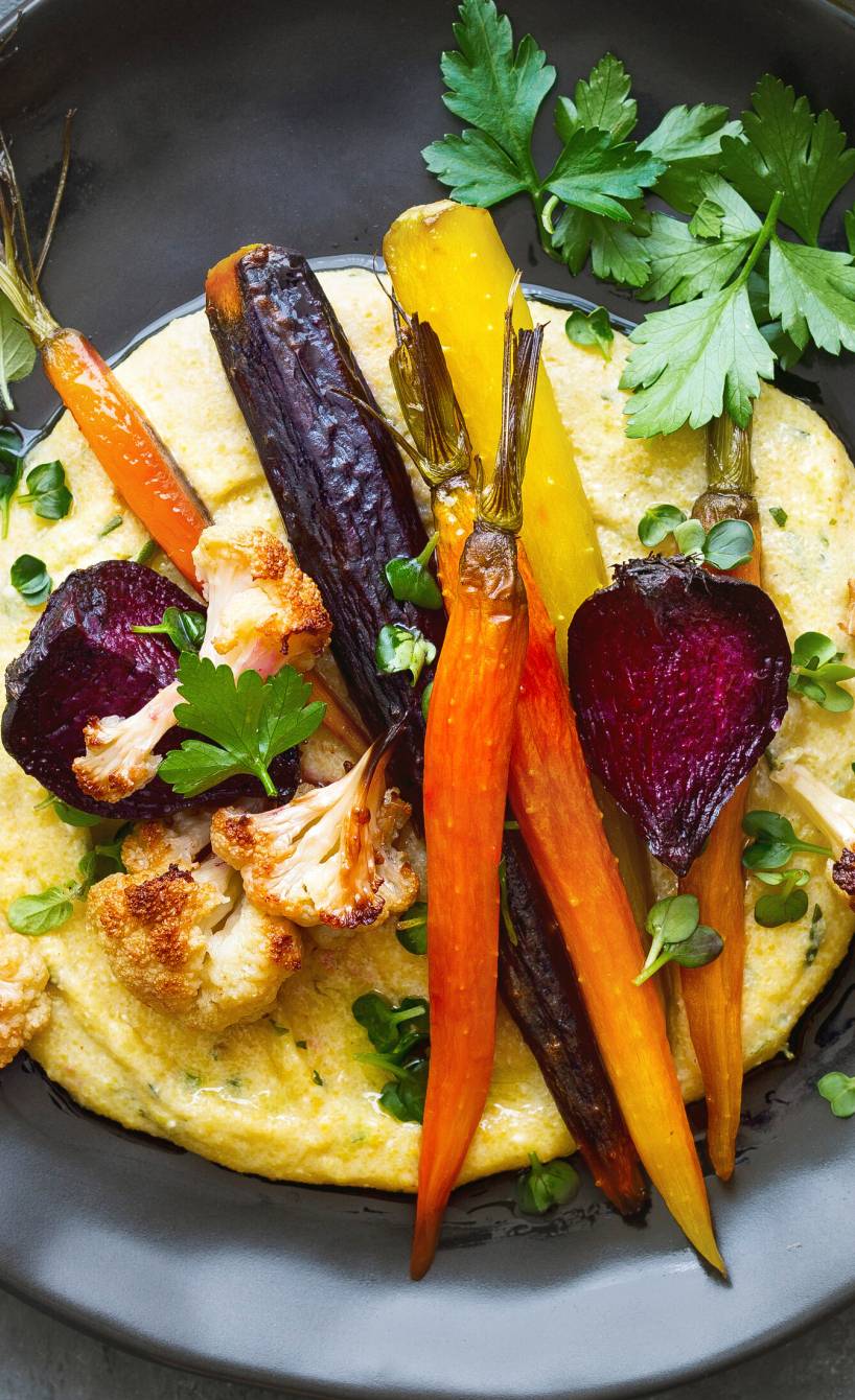 Cremige Polenta auf einem Teller getoppt von Gemüse aus dem Ofen, darauf frische Kräuter