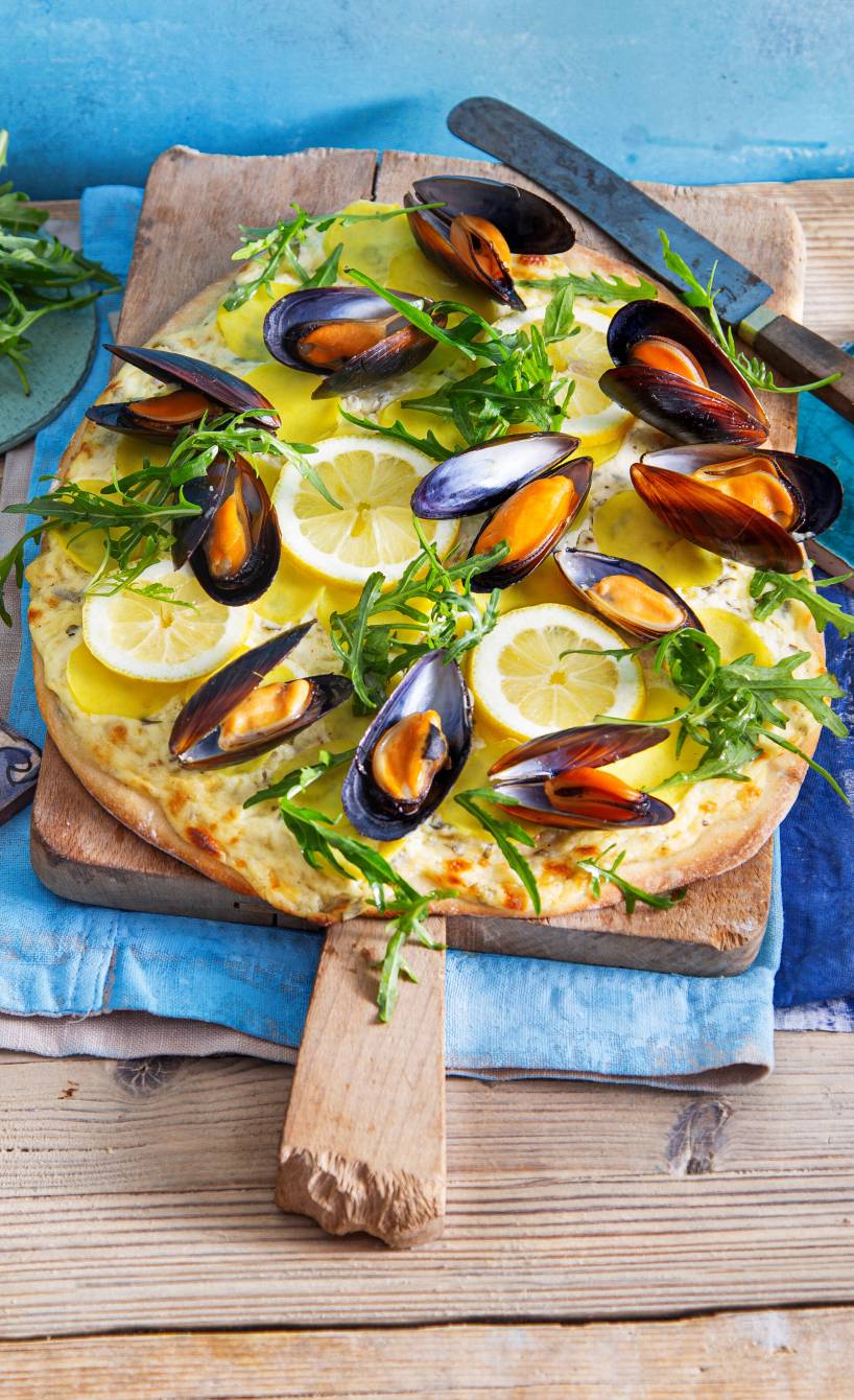 Ovale Pizza Bianca mit Miesmuscheln, Kartoffeln und Rucola auf einem Holzbrett garniert mit Zitronenscheiben