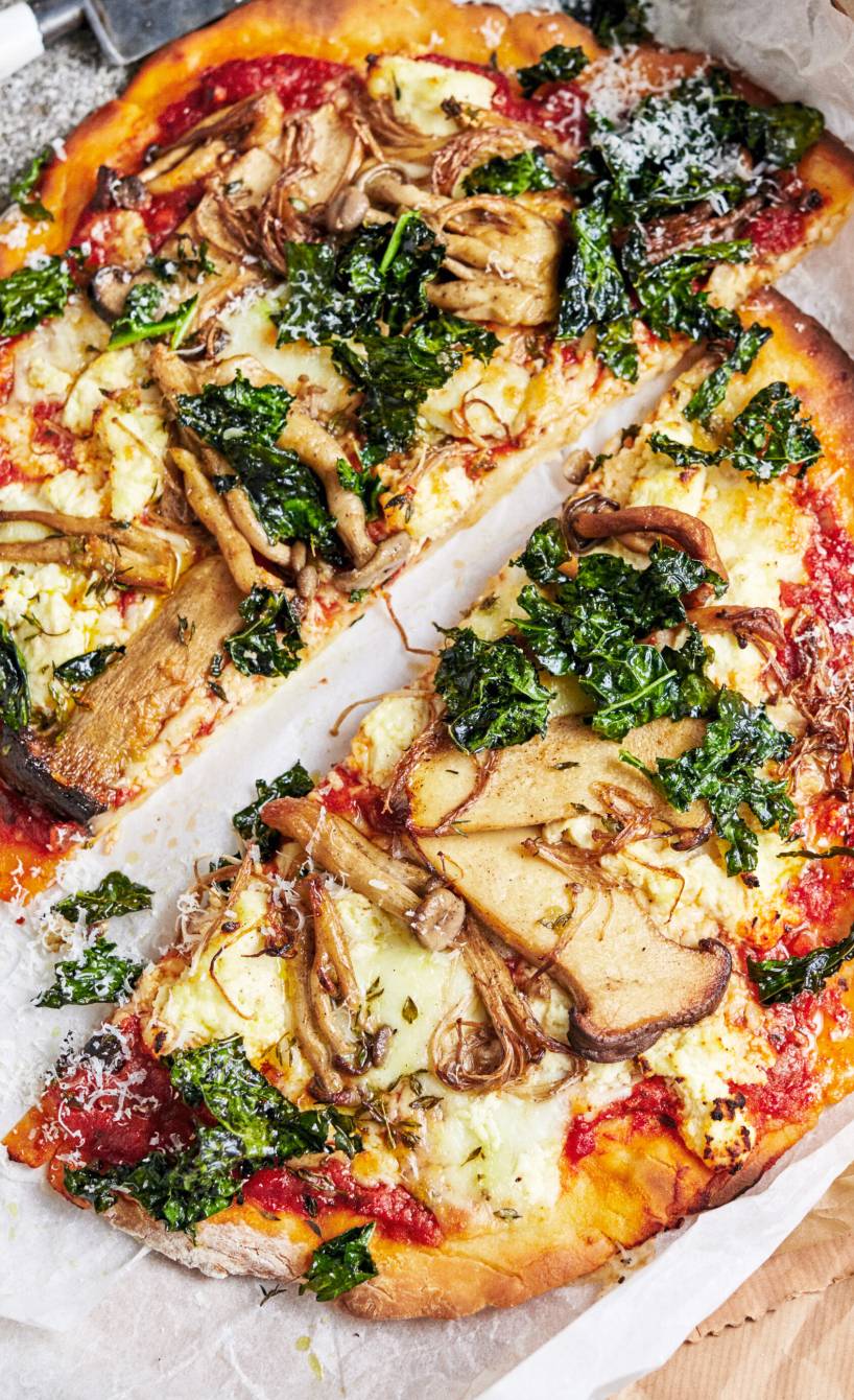 Runde Sauerteigpizza mit Pilzen, Schwarzkohl und dreierlei Käse belegt halbiert auf einem Holzbrett