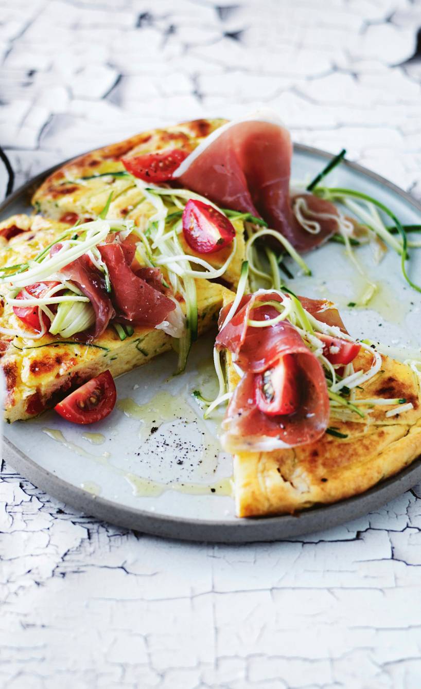 Runde Zucchini-Frittata aus Polenta angeschnitten auf einem Teller getoppt von Kirschtomaten und rohem Schinken
