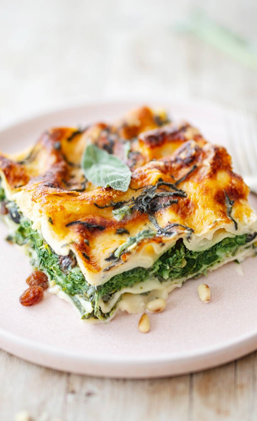 Spinatlasagnestück auf einem rosa Teller, daneben ein paar Rosinen