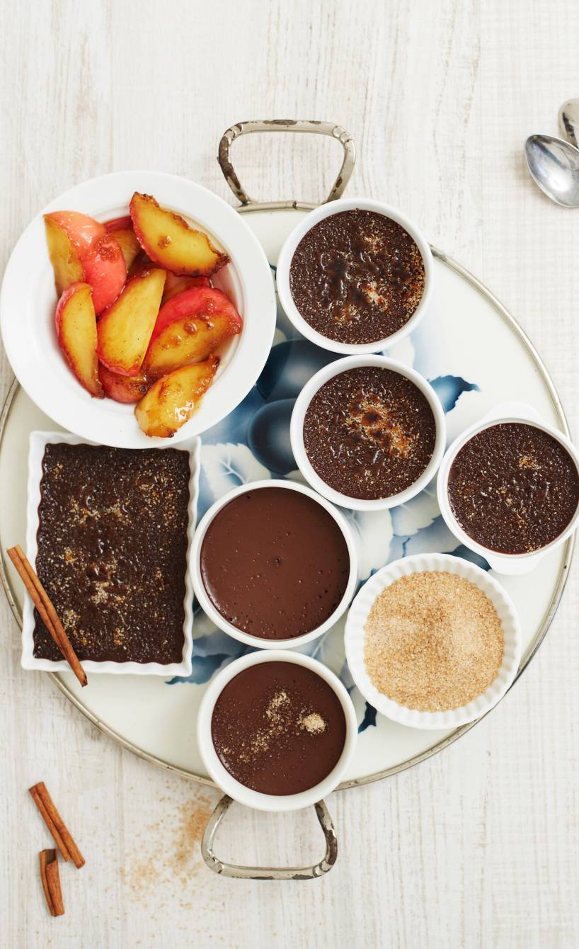 Fünf Ofenförmchen mit Schokoladen-Crème-Brûlée auf einem Tablett, daneben eine Schale mit karamellisierten Äpfeln