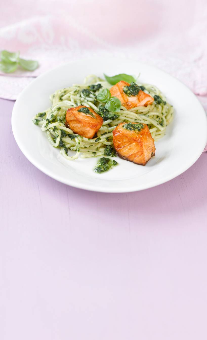Spaghetti auf einem Teller getoppt von grünem Pesto und gebratenen Lachsstücken, daneben Zitronenviertel