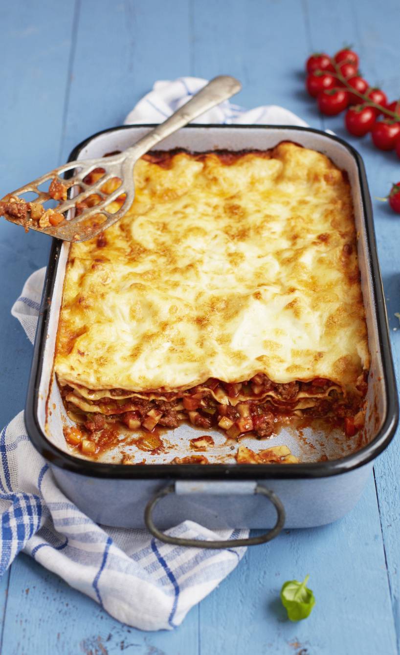 Klassische Lasagne mit Bolognese auf blauem Untergrund und Geschirrtuch, Käse und Kirschtomaten im Hintergrund, ein Portionierlöffel liegt auf der Form, Basilikumblättchen im Vordergrund