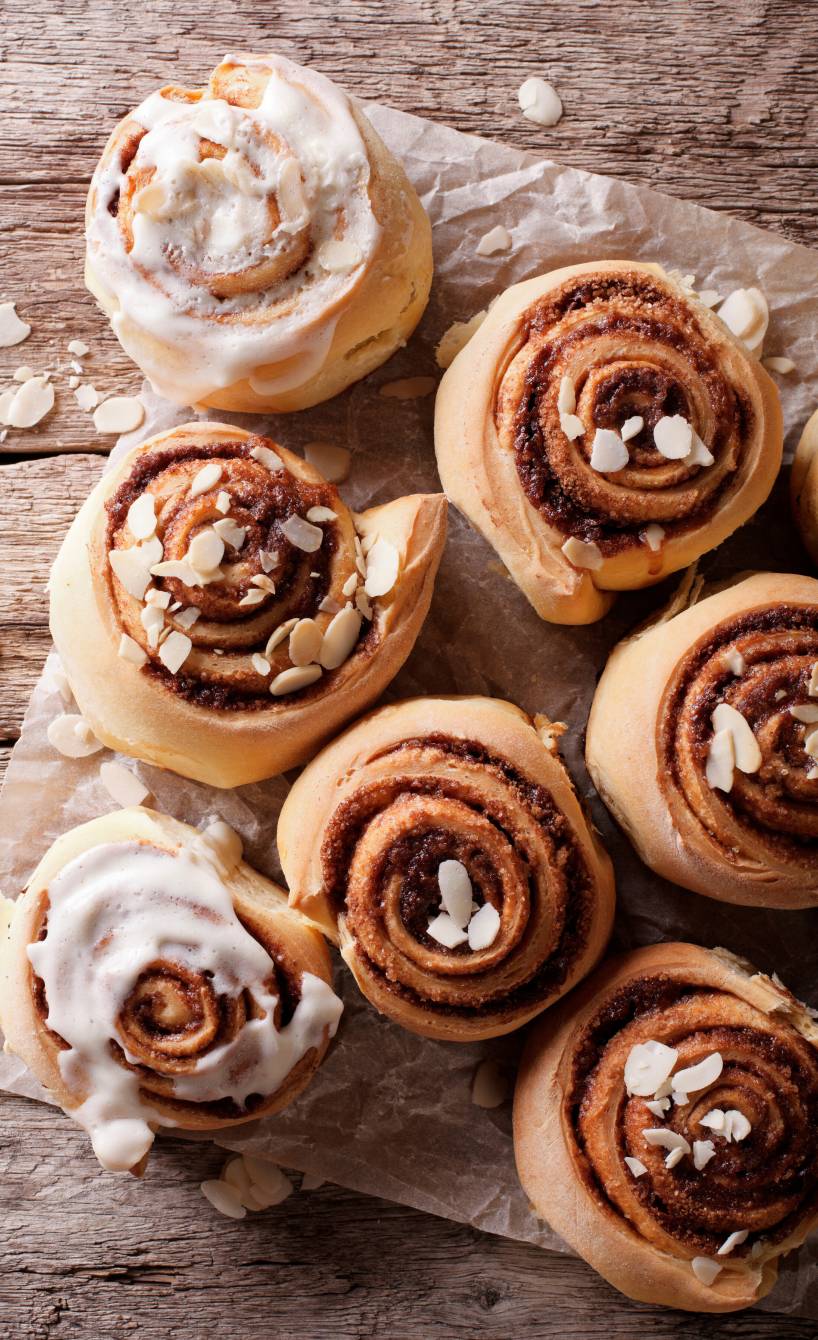 Mehrere selbst gebackene Mandelschnecken aus Hefeteig mit Zimt auf Backpapier, einige mit Glasur und Mandelblätchen verziert