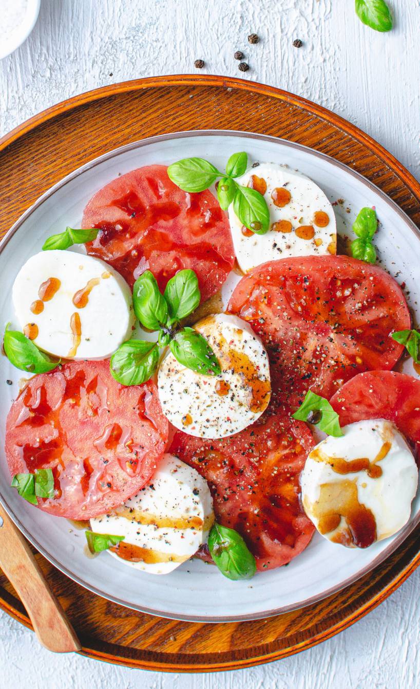 Tomaten- und Mozzarellascheiben auf einem Holzteller angerichtet als Caprese-Salat getoppt mit Balsamicodressing