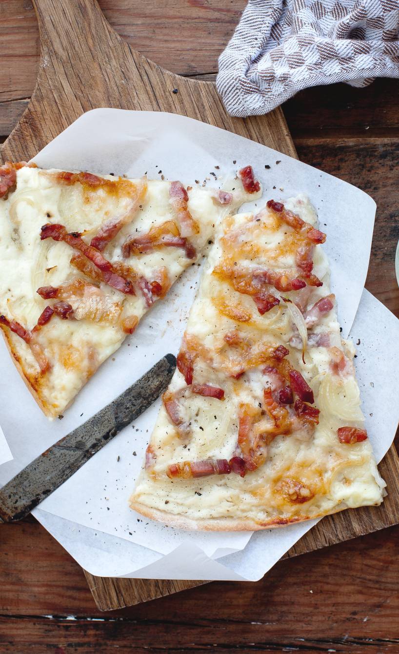 Elsässer Flammkuchen in Pizzastücke geschnitten auf einem Teller, daneben Crème fraiche in einer Schüssel