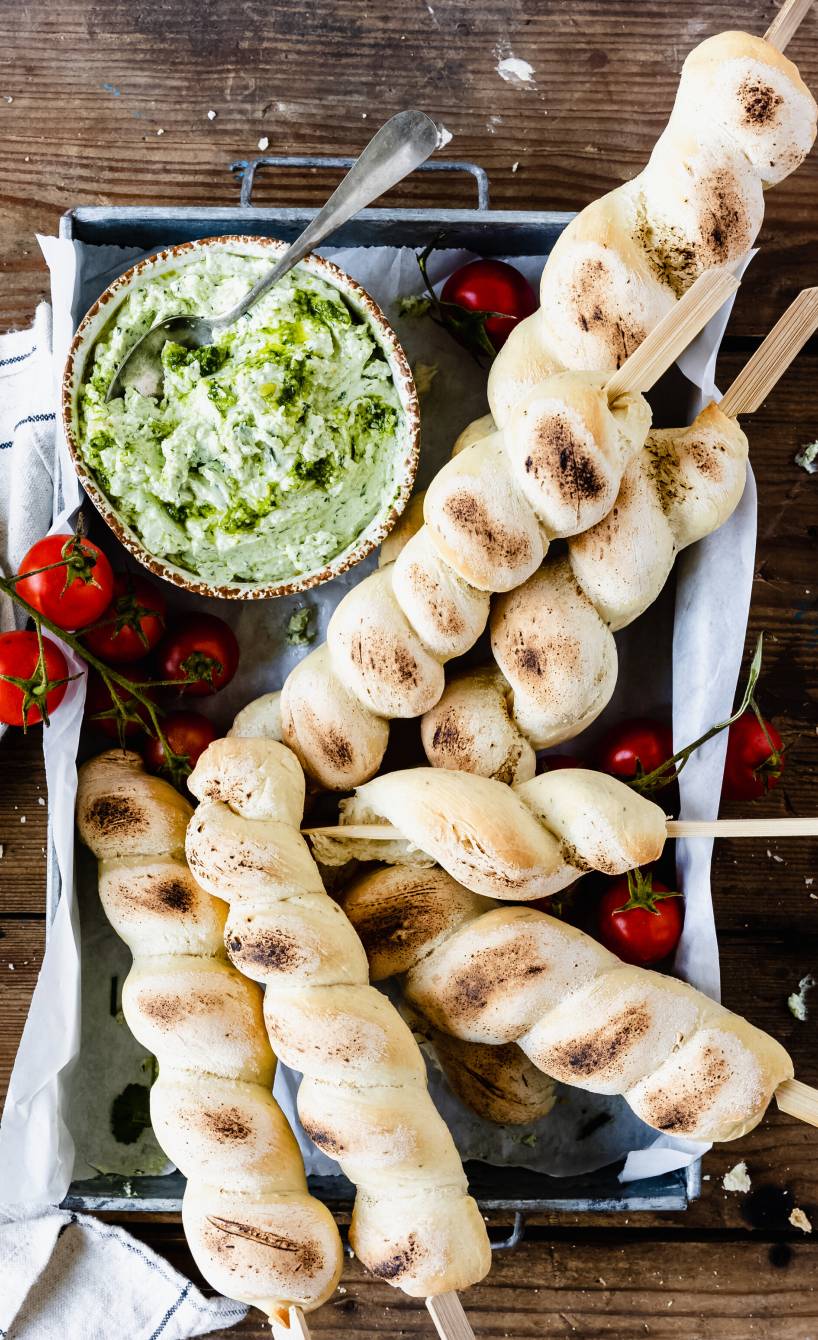 Fünf gebackene Stockbrot auf einem Backblech auf einem Holztisch, daneben Dip und Strauchtomaten