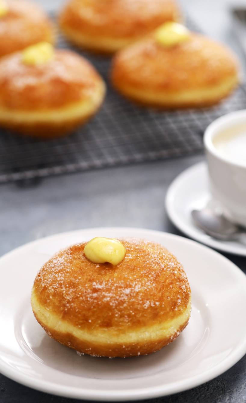Mehrere Bomboloni auf einem Kuchengitter, davor eines mit Creme gefüllt auf einem Teller