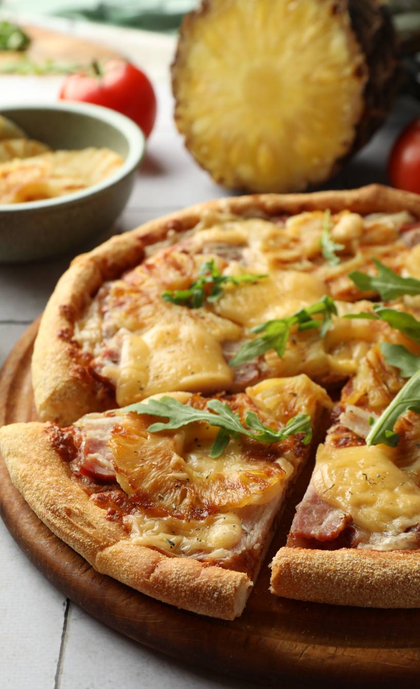 Runde Pizza Hawaii angeschnitten auf einem Holzbrett, dahinter eine halbierte Ananas
