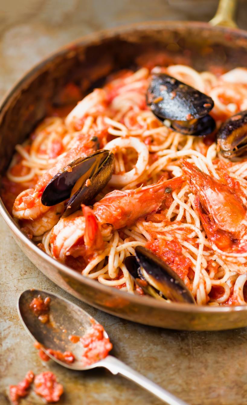 Spaghetti Frutti di Mare in einer Pfanne, daneben ein Löffel mit Tomatensauce