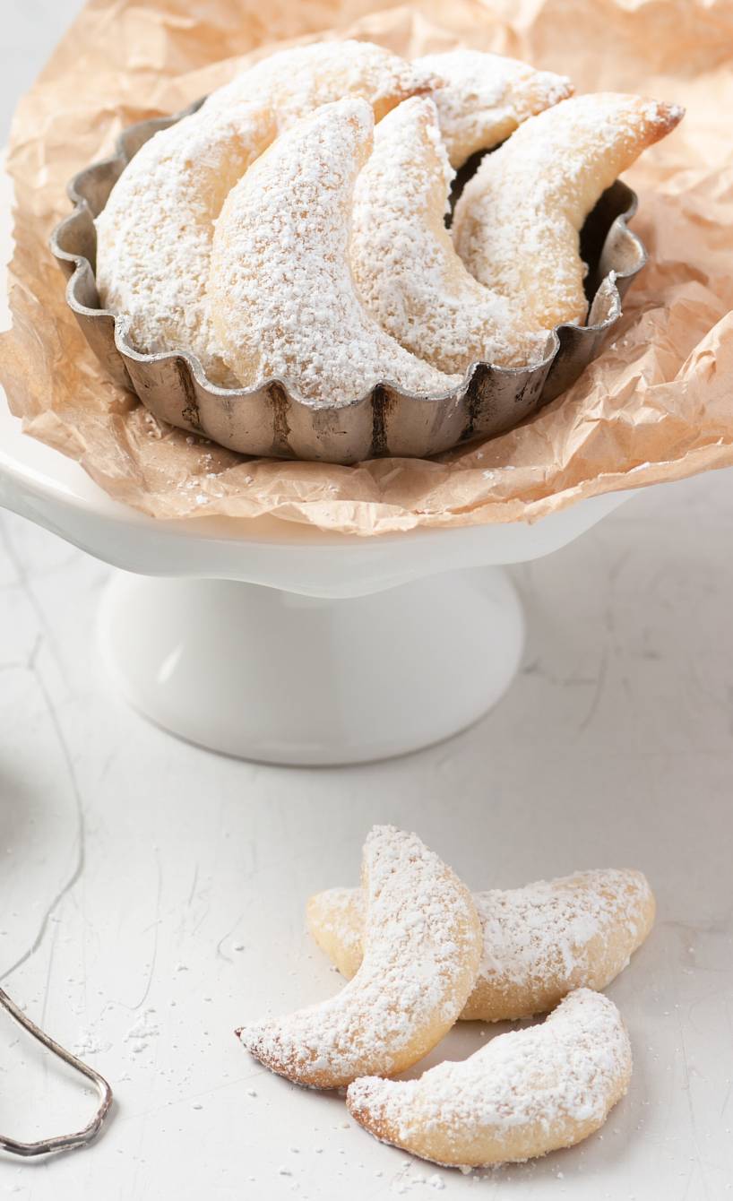 Vanillekipferl auf einer Tortenplatte, daneben Puderzucker auf einem Tisch