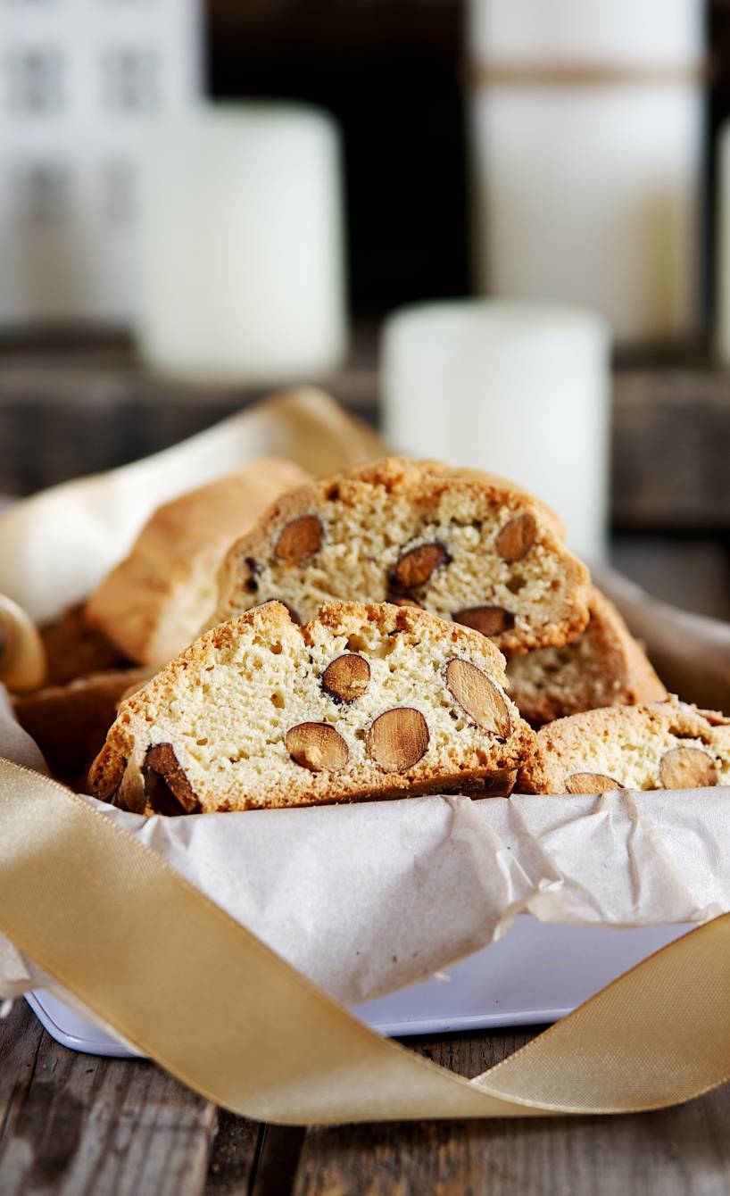 Selbstgemachte Cantuccini in einer Geschenkschachtel auf einem Tisch, dahinter Weihnachtsdeko