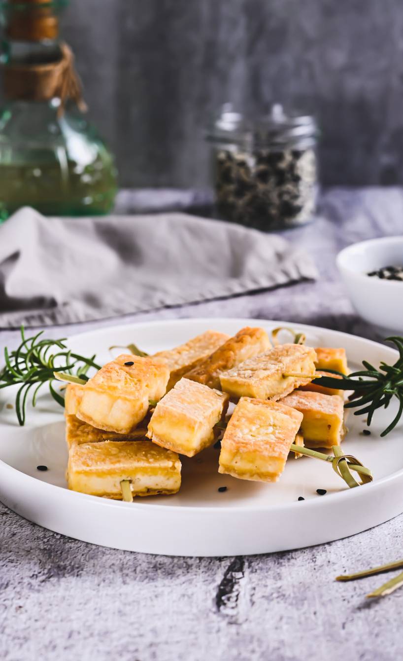 Gegrillter Tofu auf Spießen auf einem Teller, daneben Rosmarinzweige, Olivenöl und ein Glas schwarzer Pfeffer
