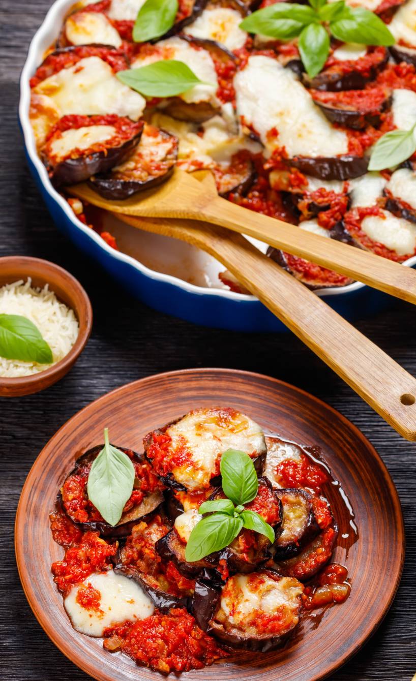Veganer Auberginen-Auflauf Parmigiana in einer runden Auflaufform, daneben eine Portion auf einem Teller sowie frischer Basilkum