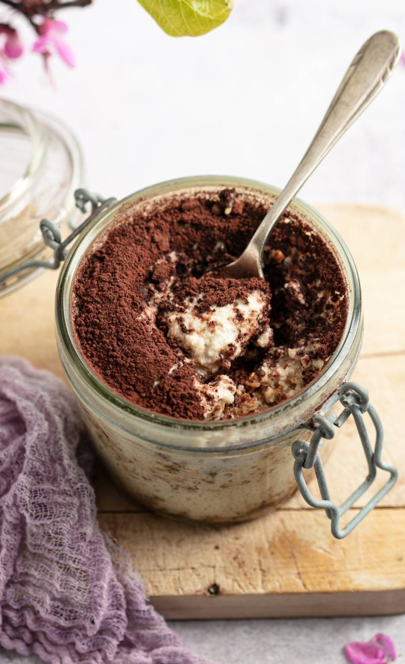 Veganes Tiramisu in einem geöffneten Weckglas auf einem Holzbrett, darin ein Dessertlöffel