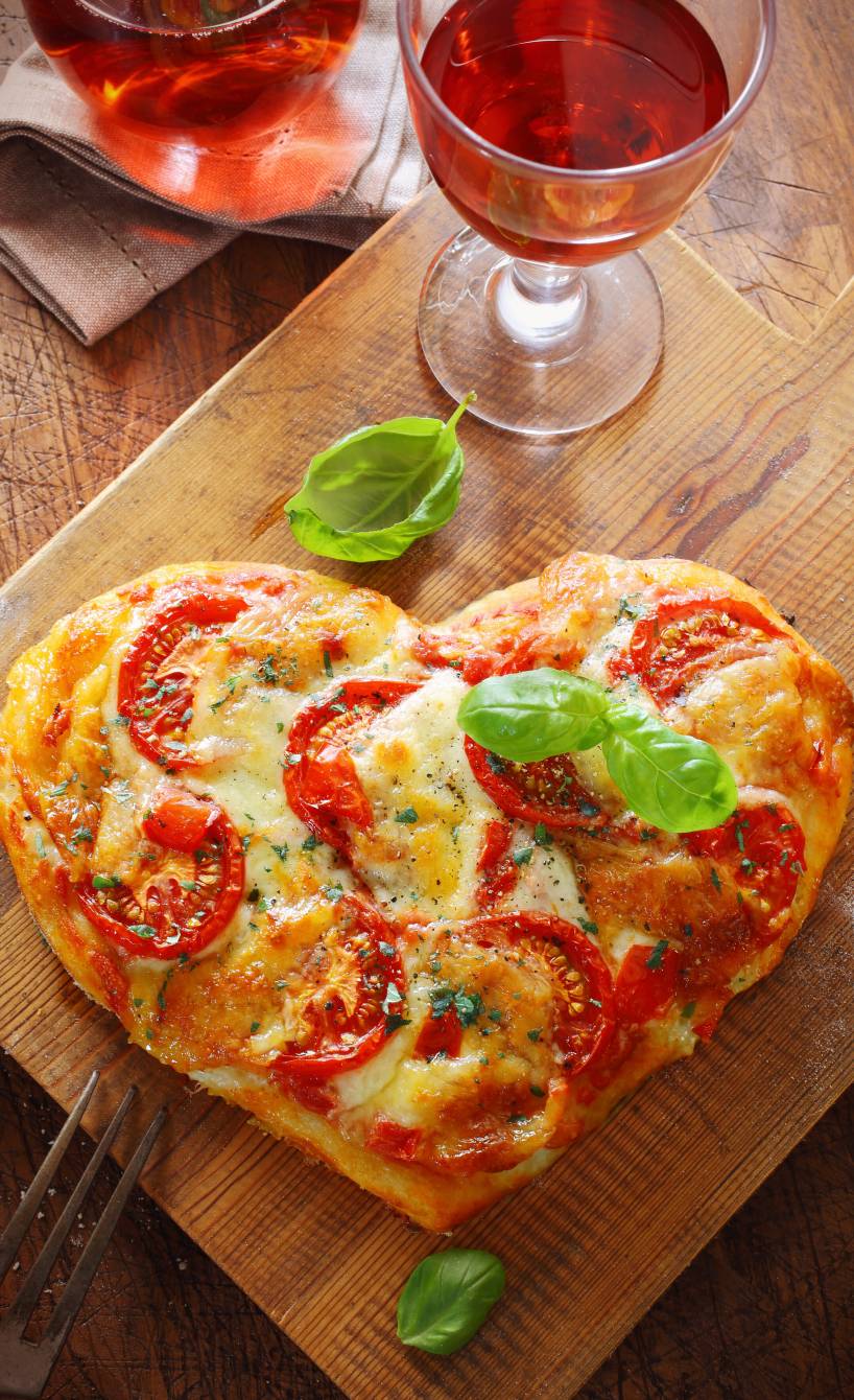 Pizza in Herzform auf einem Holztisch garniert mit Oregano und Basilikum, daneben ein Glas Wein