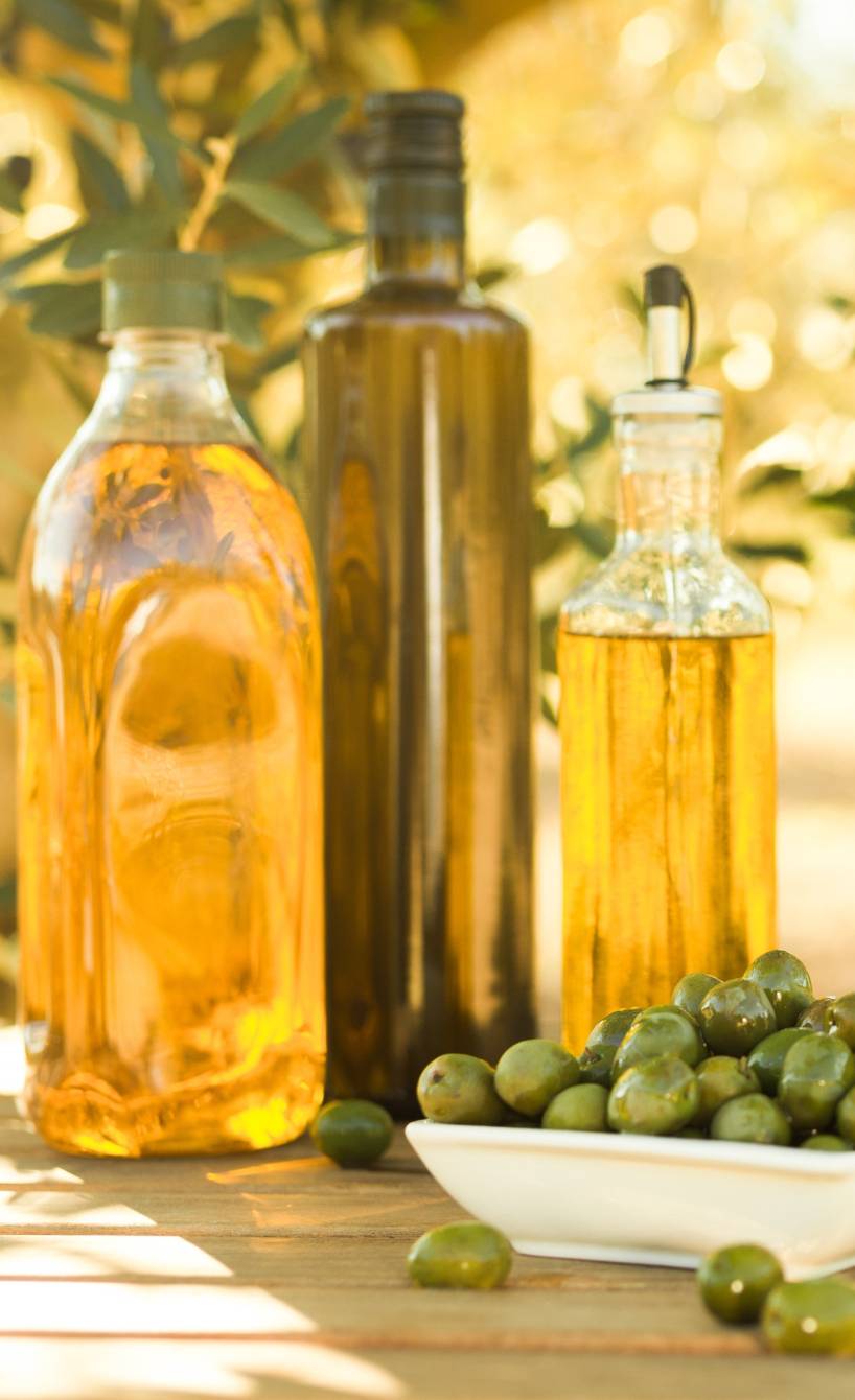 Mehrere Flaschen Olivenöl sowie grüne Oliven auf einem Holztisch vor einem Olivenhain in Italien