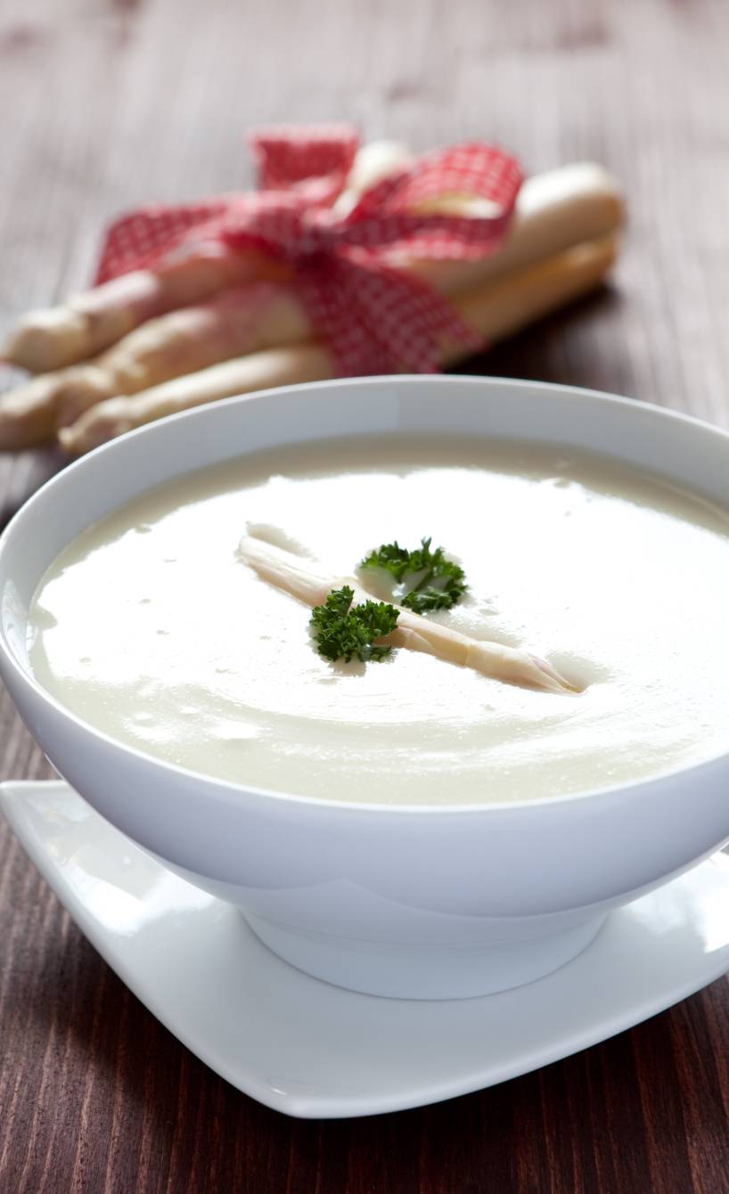 Spargelcremesuppe in einer Suppenschüssel, dahinter weißer Spargel
