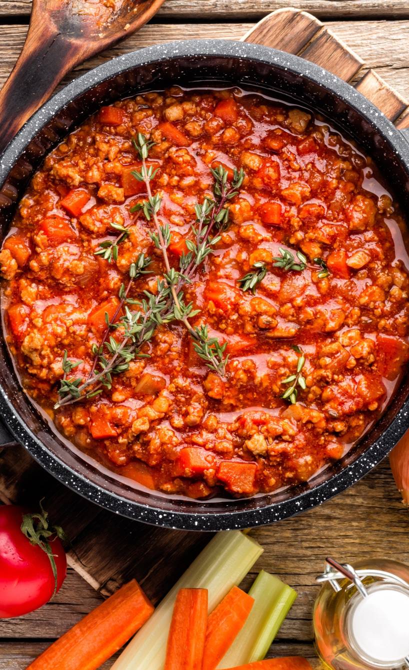 Frisch gekochte Bolognese auf einem Holztisch, daneben Zutaten wie Gemüse und Kräuter