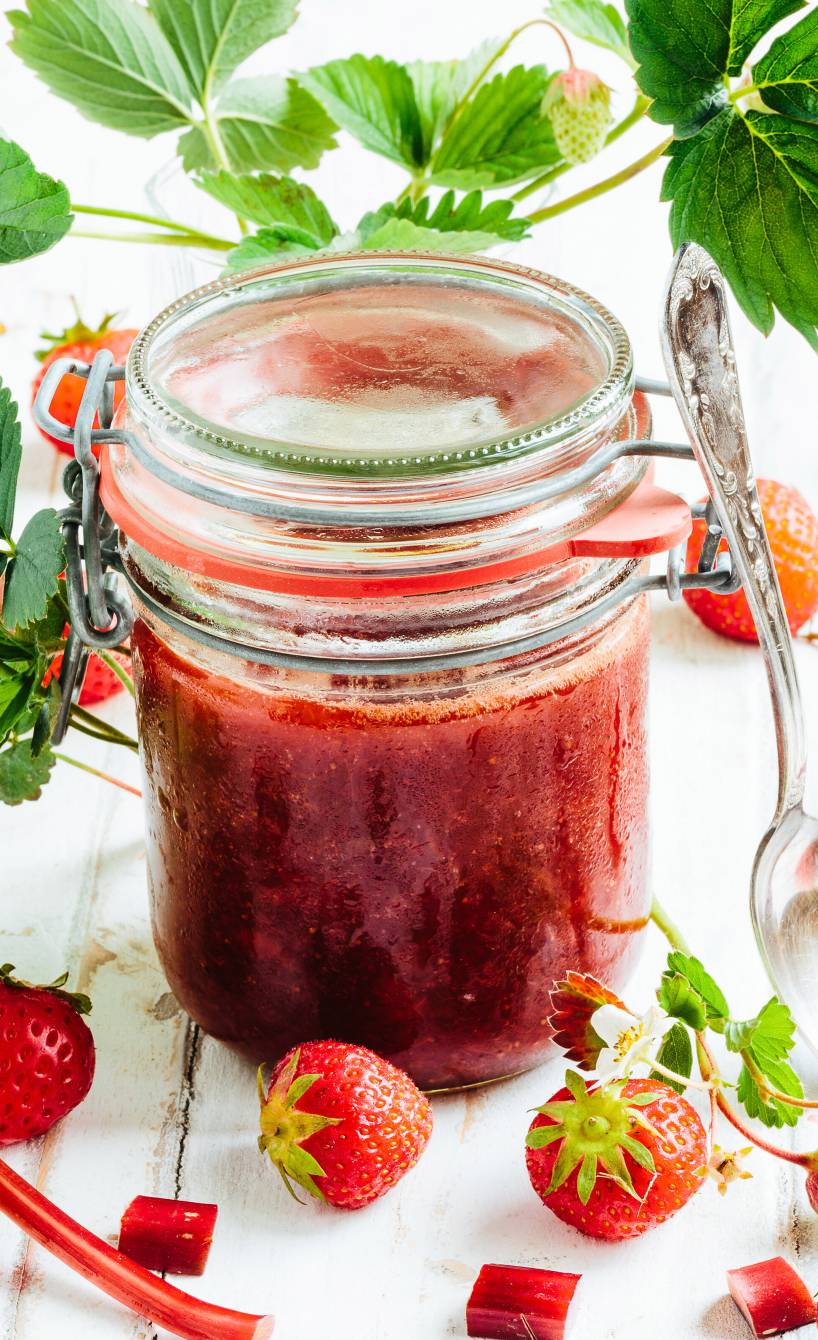Erdbeer-Rhabarber-Marmelade in einem geschlossen Weckglas auf einem Holztisch, daneben Erdbeeren und Rabarberstücke