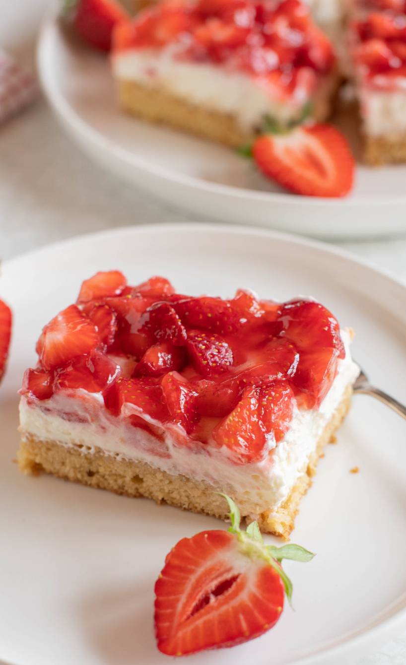Ein Stück Erdbeerkuchen mit Pudding-Quark-Creme, dahinter das Blech mit dem restlichen Blechkuchen