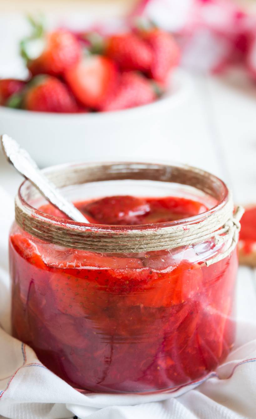 Selbst gemachte Erdbeermarmelade in einem Einmachglas, dahinter frische Erdbeeren auf einem Holztisch