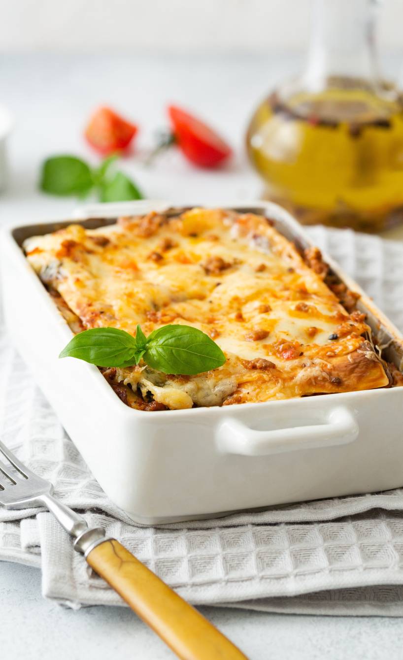 Gemüselasagne in einer Auflaufform auf einem Tisch, dahinter Olivenöl und Tomaten