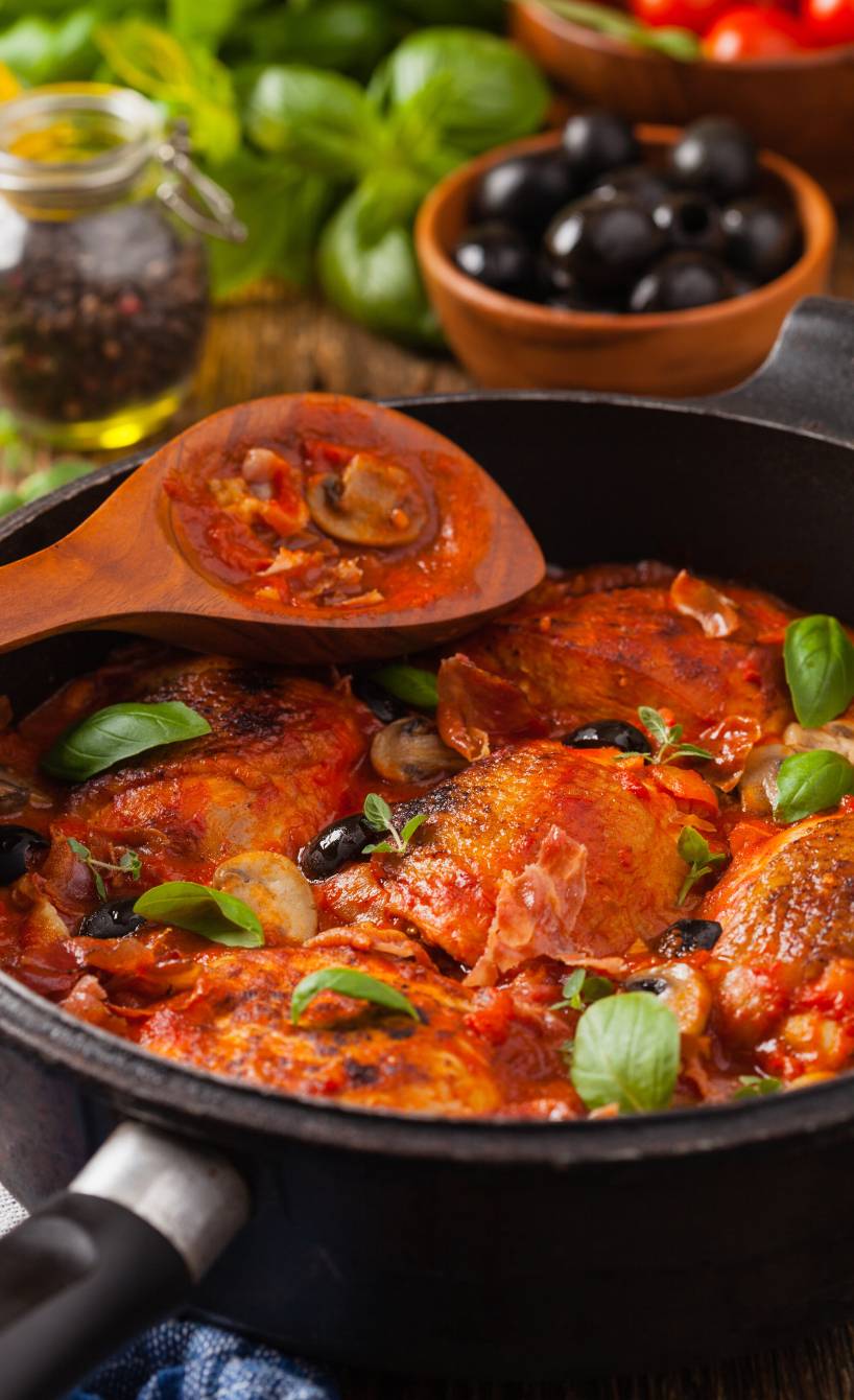 Geschmorte Hähnchenschenkel alla cacciatora in Tomatensauce in einer Pfanne garniert mit schwarzen Oliven und frischen Kräutern